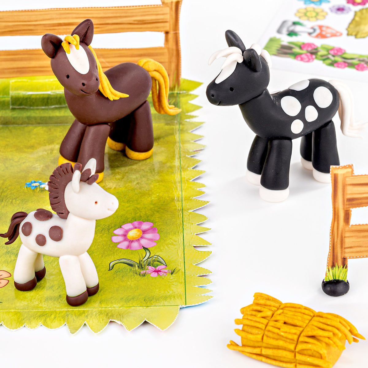 Finished FIMO kids horses in brown, black with white spots and white with brown spots, standing on the meadow playing surface by a wooden fence