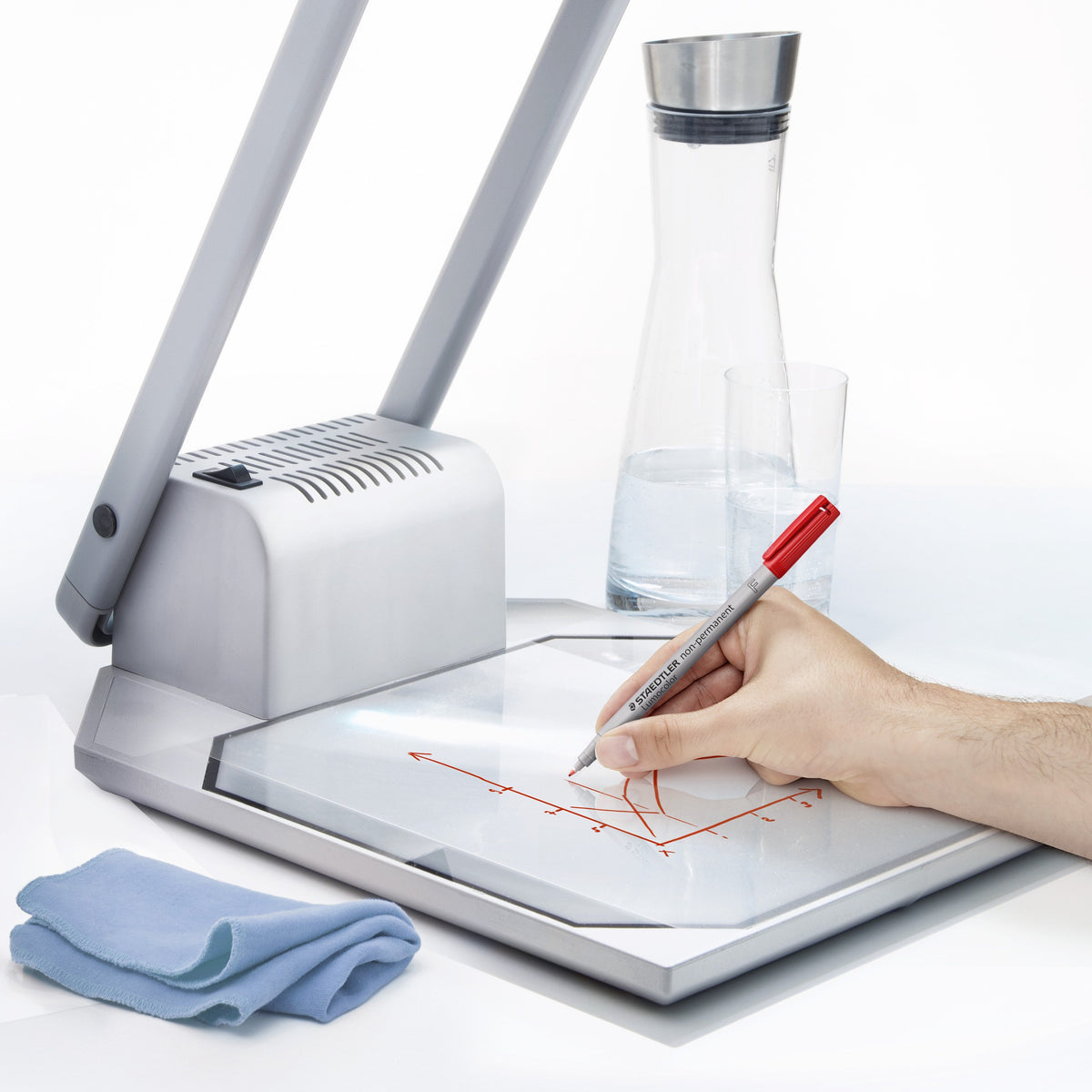 Hand writing on overhead film with a red Lumocolor non-permanent pen beside a carafe of water and a blue cloth