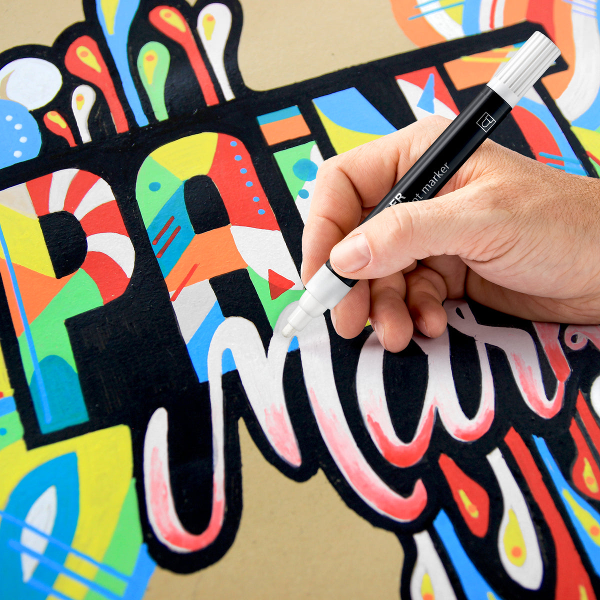 Painted lettering on brown card being detailed by hand with a white capped Lumocolor paint marker from the same 349 range