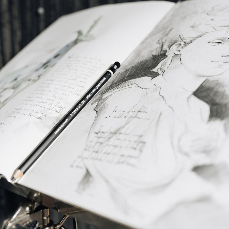 Black Mars Lumograph black pencil resting on an open sketchbook beside a soft pencil portrait, Staedtler mood photograph