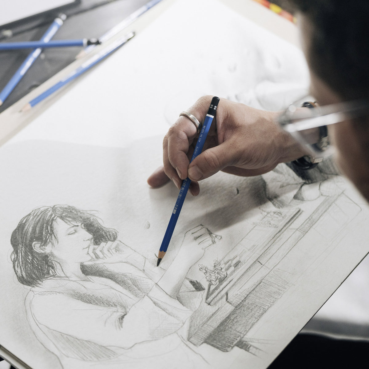 Artist shading a pencil portrait of a woman in a sketchbook with a blue Mars Lumograph pencil, range photo from Staedtler