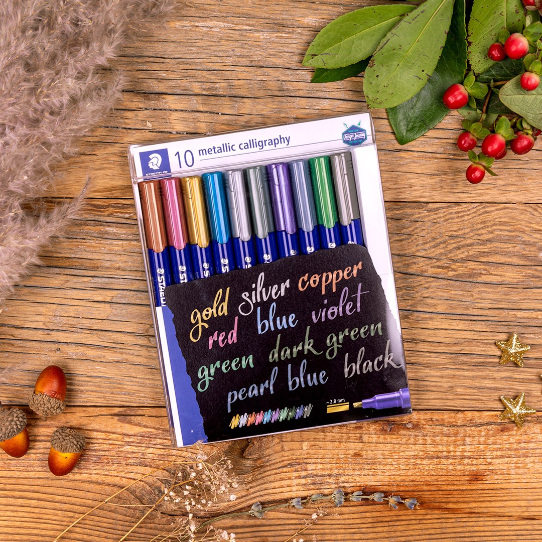 Staedtler photo of the 8325 metallic calligraphy box of 10 on a timber table with dried grass, acorns and berries, colour names lettered on the card