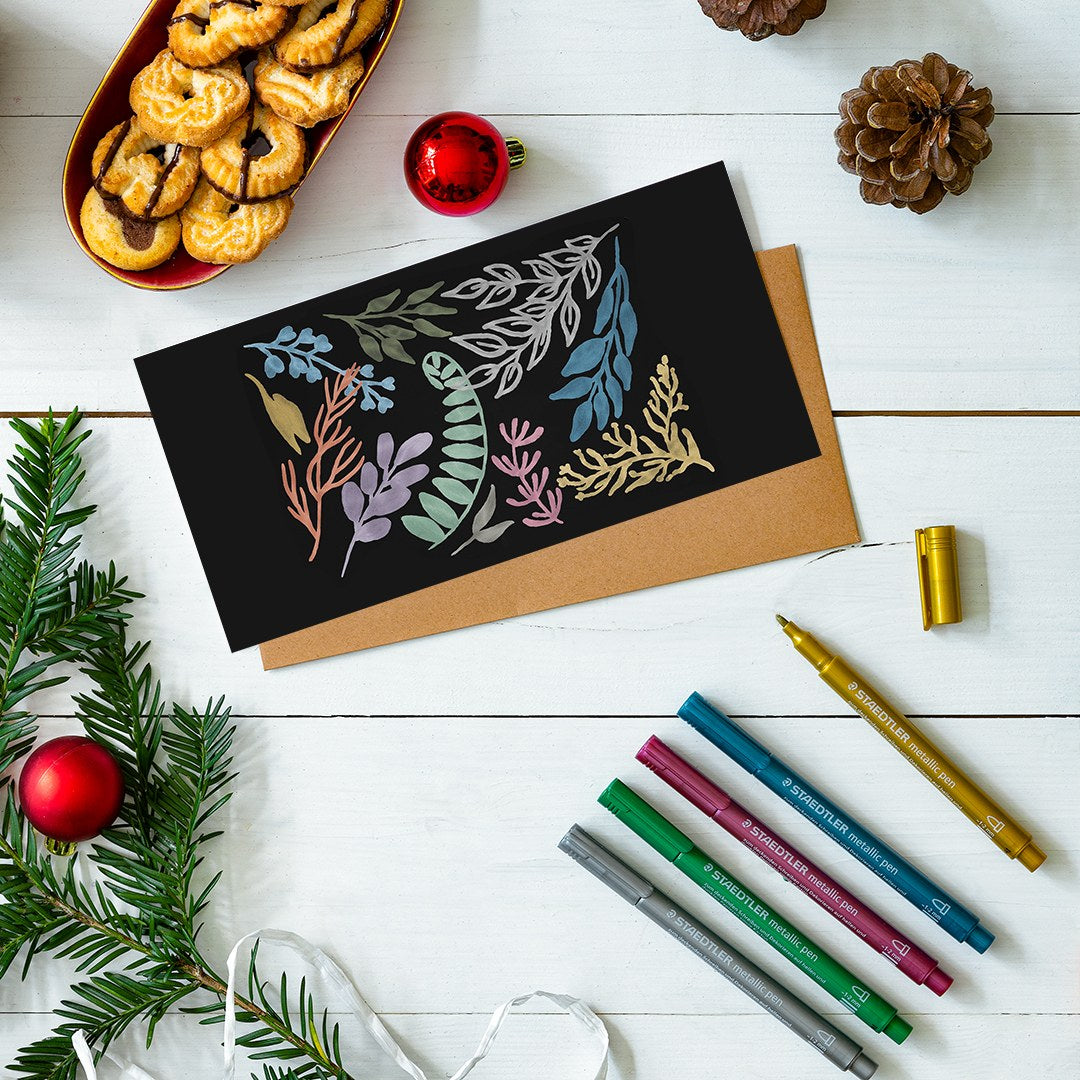 Flat lay of a black card with metallic botanical drawings on a kraft envelope, five Staedtler metallic pens, pine cones and baubles