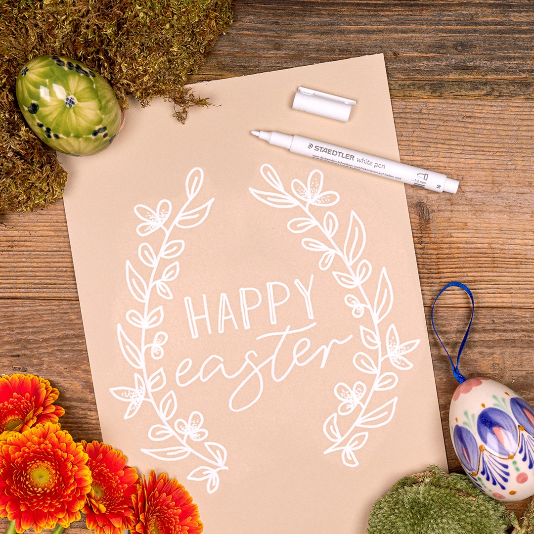 Kraft paper sheet hand lettered HAPPY easter with a white leafy wreath, a Staedtler white pen, decorated eggs and orange flowers on wood