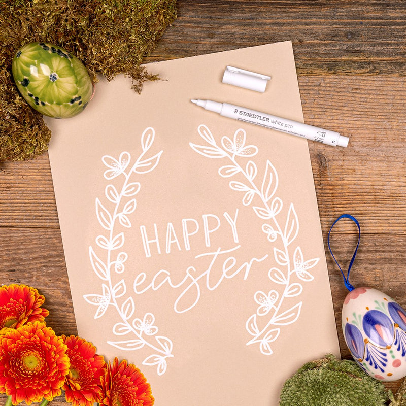 Kraft paper sheet hand lettered HAPPY easter with a white leafy wreath, a Staedtler white pen, decorated eggs and orange flowers on wood