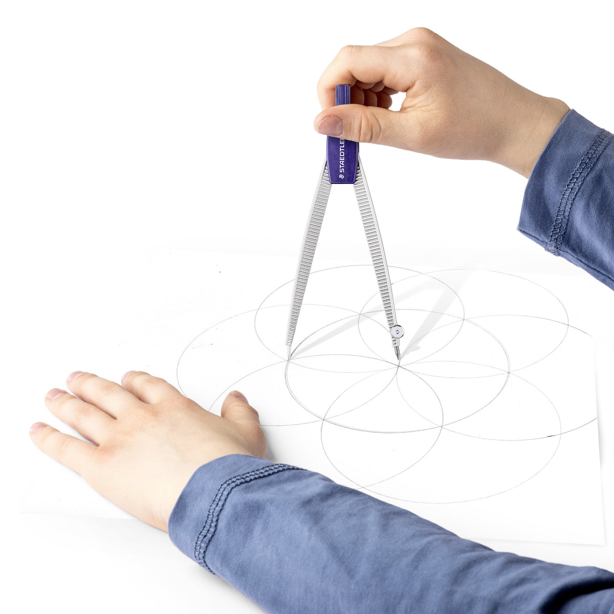 A child's hand turning a Staedtler Noris 550 compass to draw overlapping circles while the other hand steadies the paper