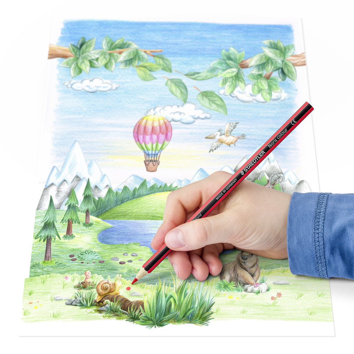 Hand colouring a landscape with mountains, a lake, trees and a hot air balloon using a red and black striped Staedtler Noris colour pencil