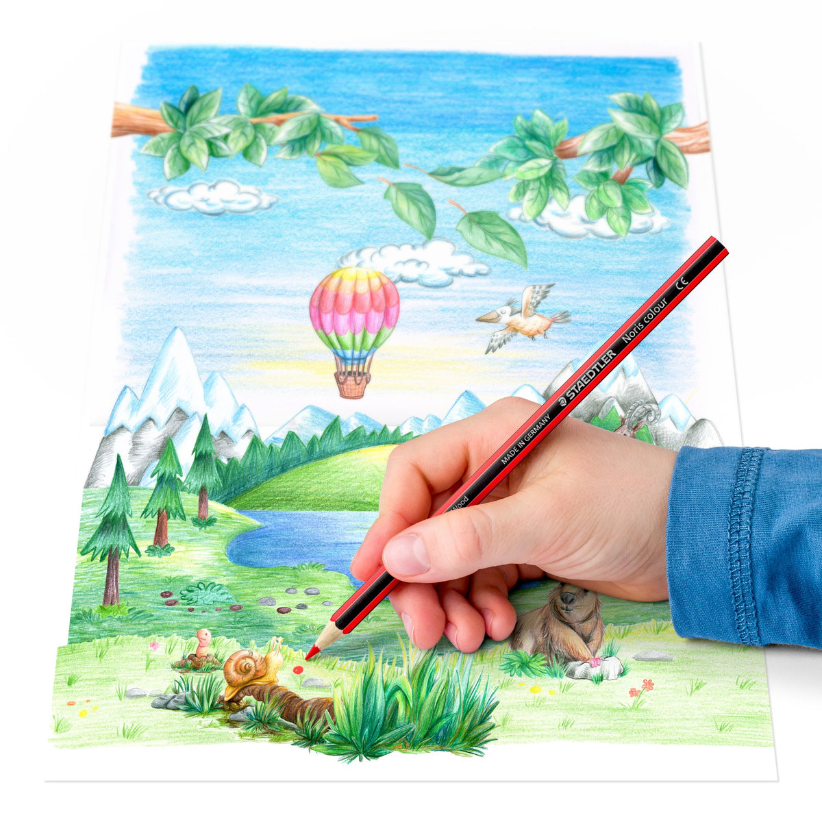 Child colouring a mountain and lake landscape with a hot air balloon using a Staedtler Noris colour pencil
