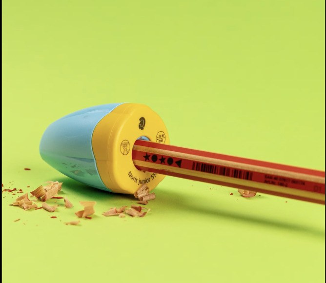 Staedtler photo of a thick red pencil in the turquoise and yellow Noris junior sharpener with shavings on a green surface