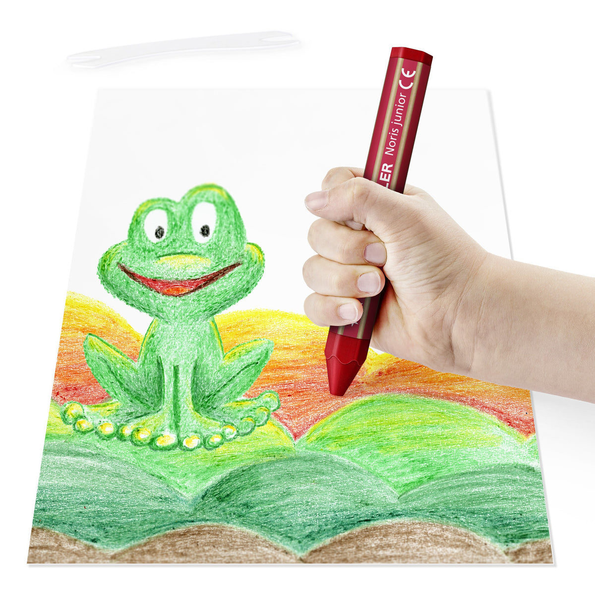 Child hand gripping a red Noris junior crayon while colouring a cheerful green frog sitting on a hill
