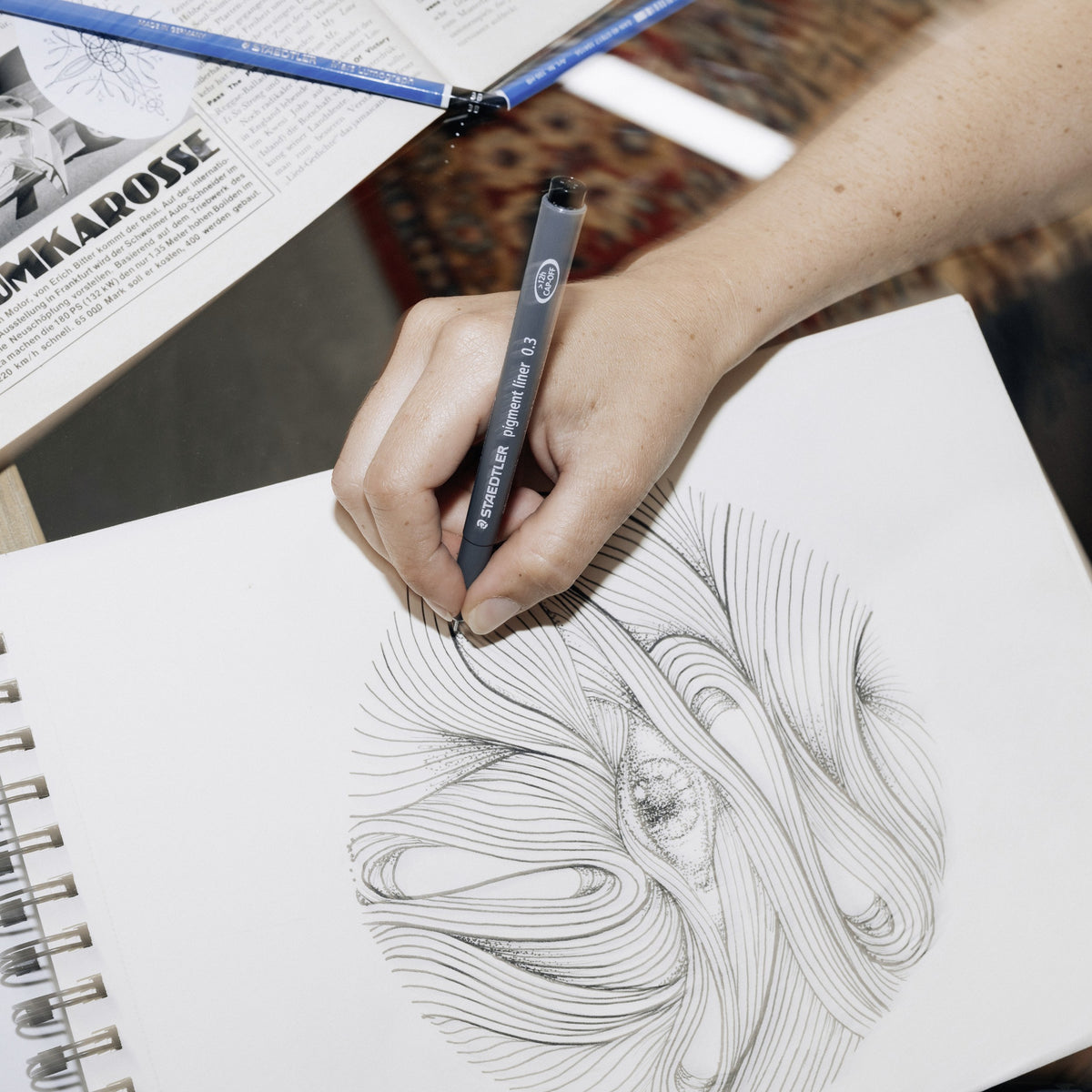 Person drawing swirling black line art in a sketchbook with a Staedtler pigment liner