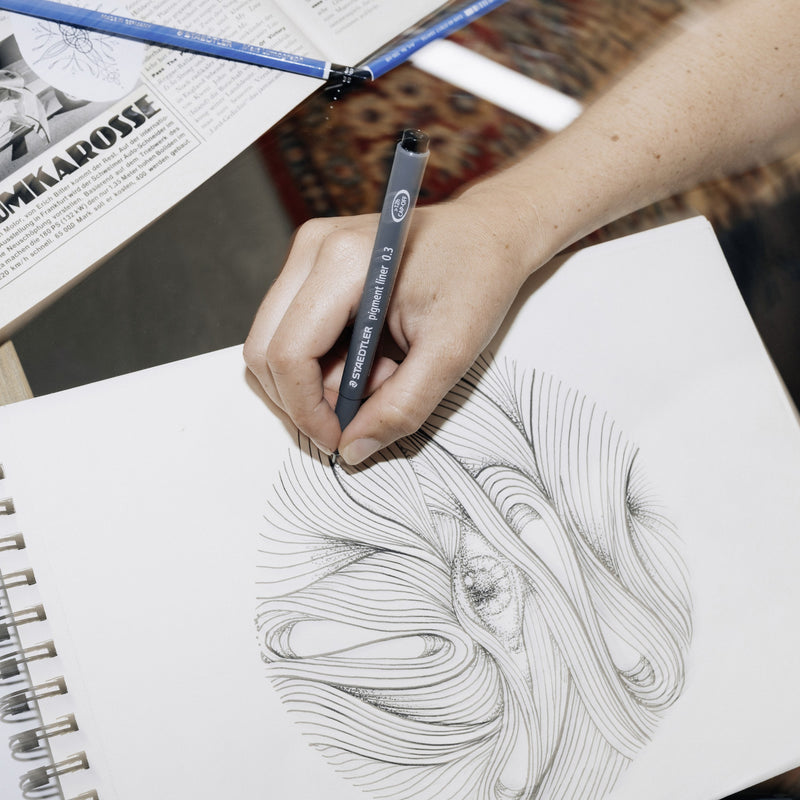 Person drawing swirling black line art in a sketchbook with a Staedtler pigment liner