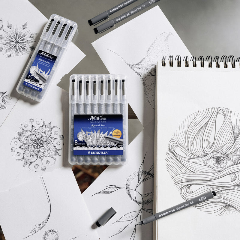 Desk scene with Staedtler pigment liner boxes of 4 and 8, loose pens and sketchbook drawings of a mandala and an eye