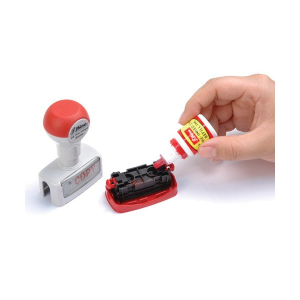 Hand squeezing drops from a small Shiny red ink bottle onto a pre-inked stamp turned face up, beside a red-handled rubber stamp