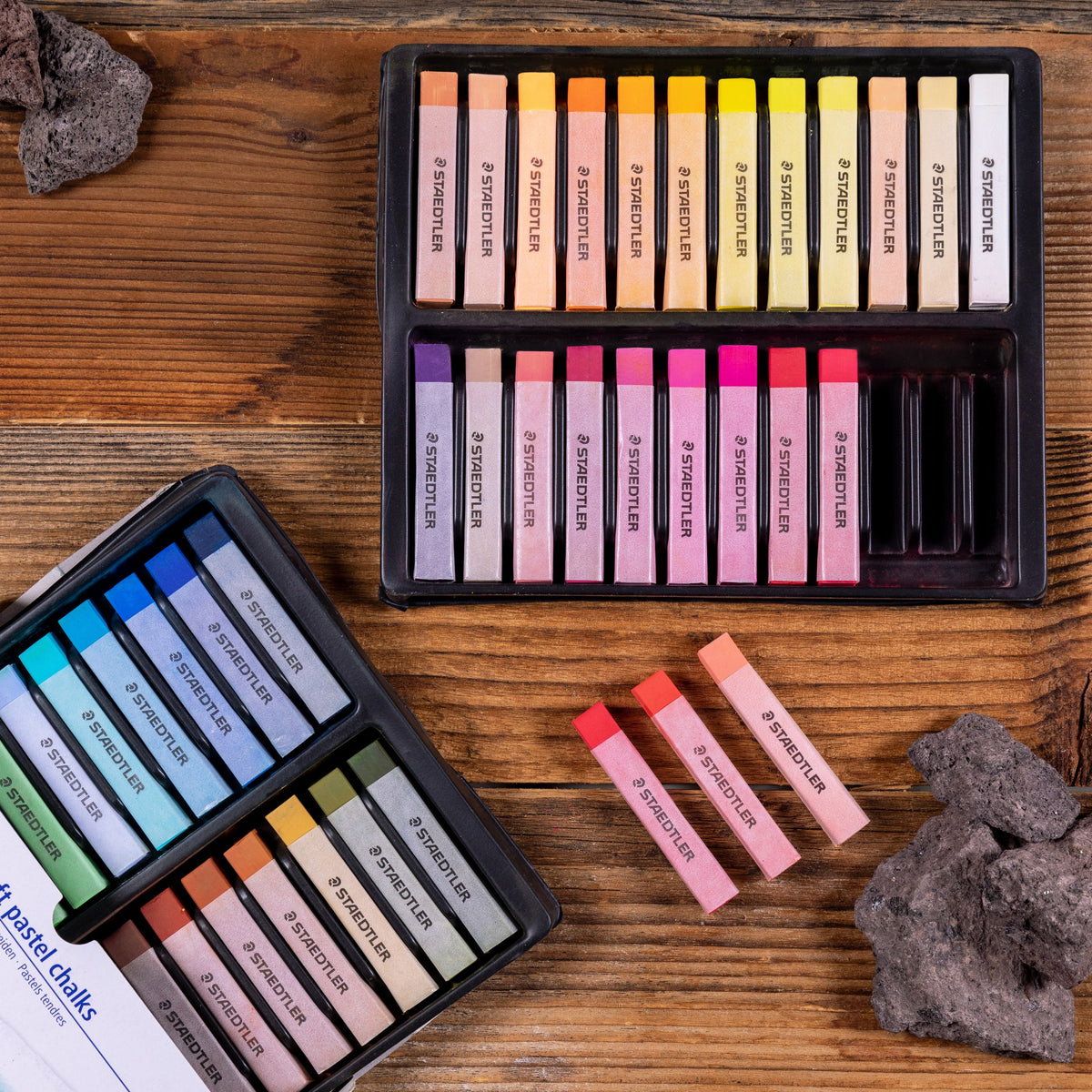 Staedtler range photo: tray of 2430 soft pastel chalks on a wooden table with loose sticks and rocks; this shows the 48 set, not the 36 box