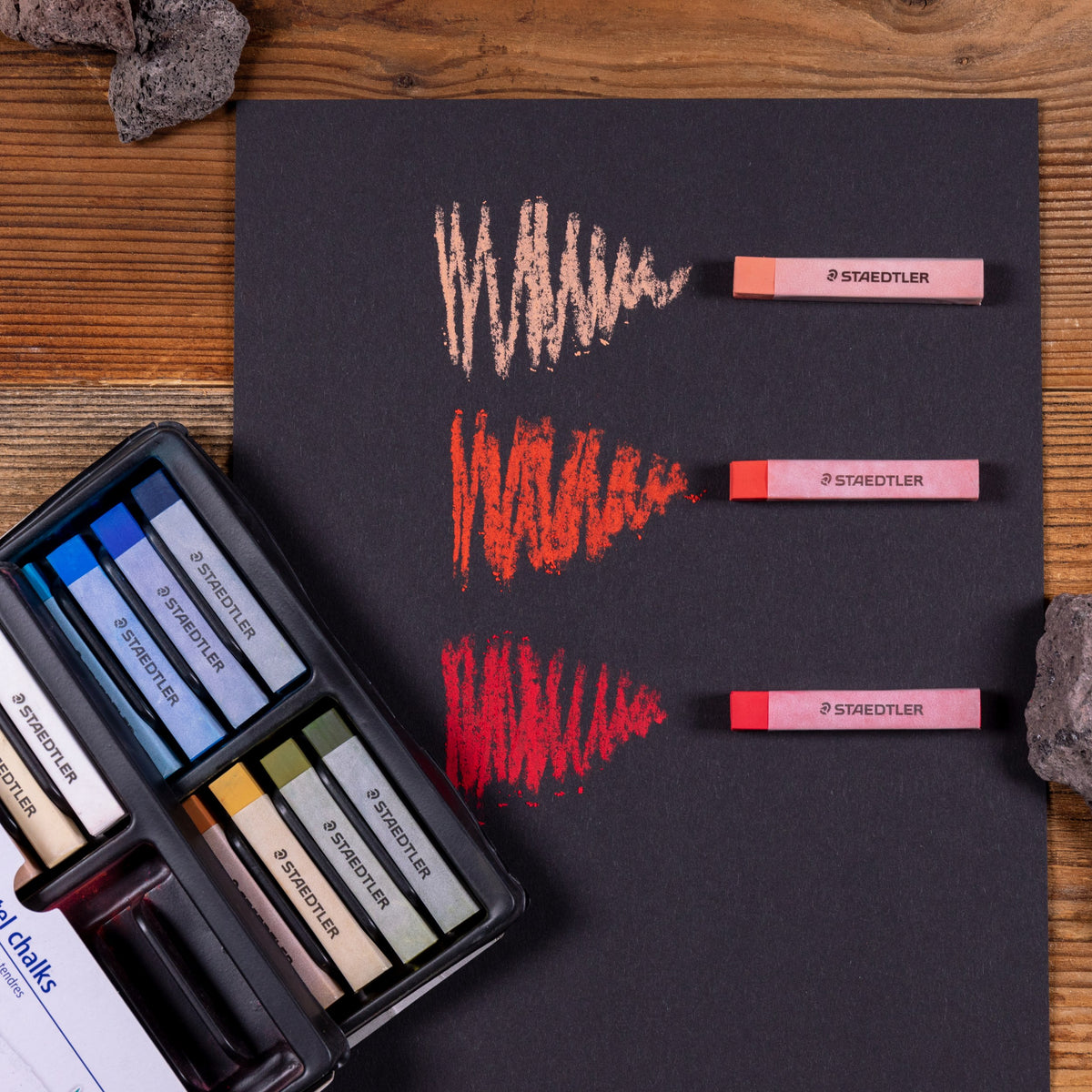 Staedtler range photo: pastel strokes in pinks and reds on black paper beside an open tray of soft pastels from the 48 set