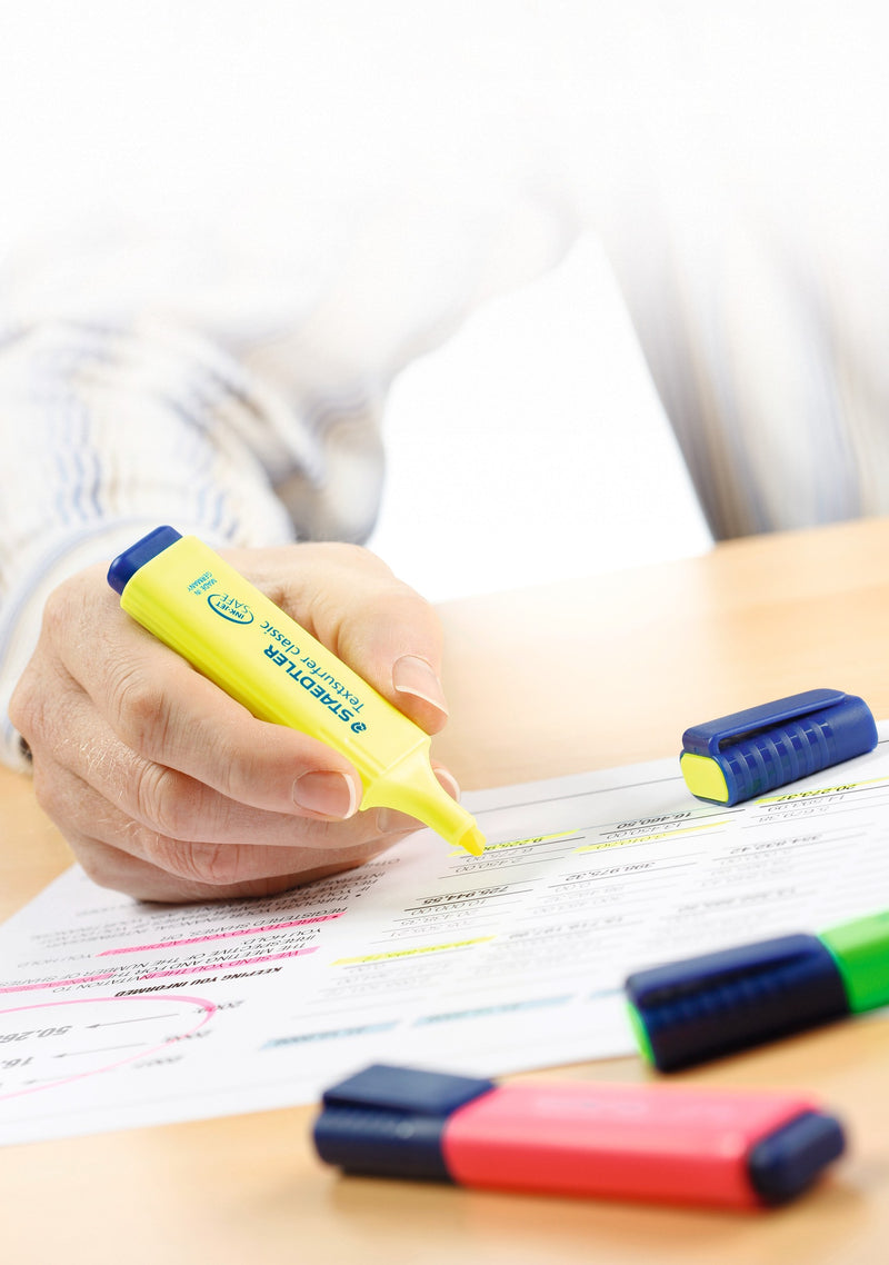 Desk scene from Staedtler with a hand highlighting figures in yellow while capped Textsurfer classic pens lie on the table