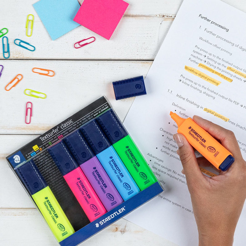 Staedtler photo of the open wallet of 6 on a desk with paper clips and notes, a hand highlighting text with the orange pen