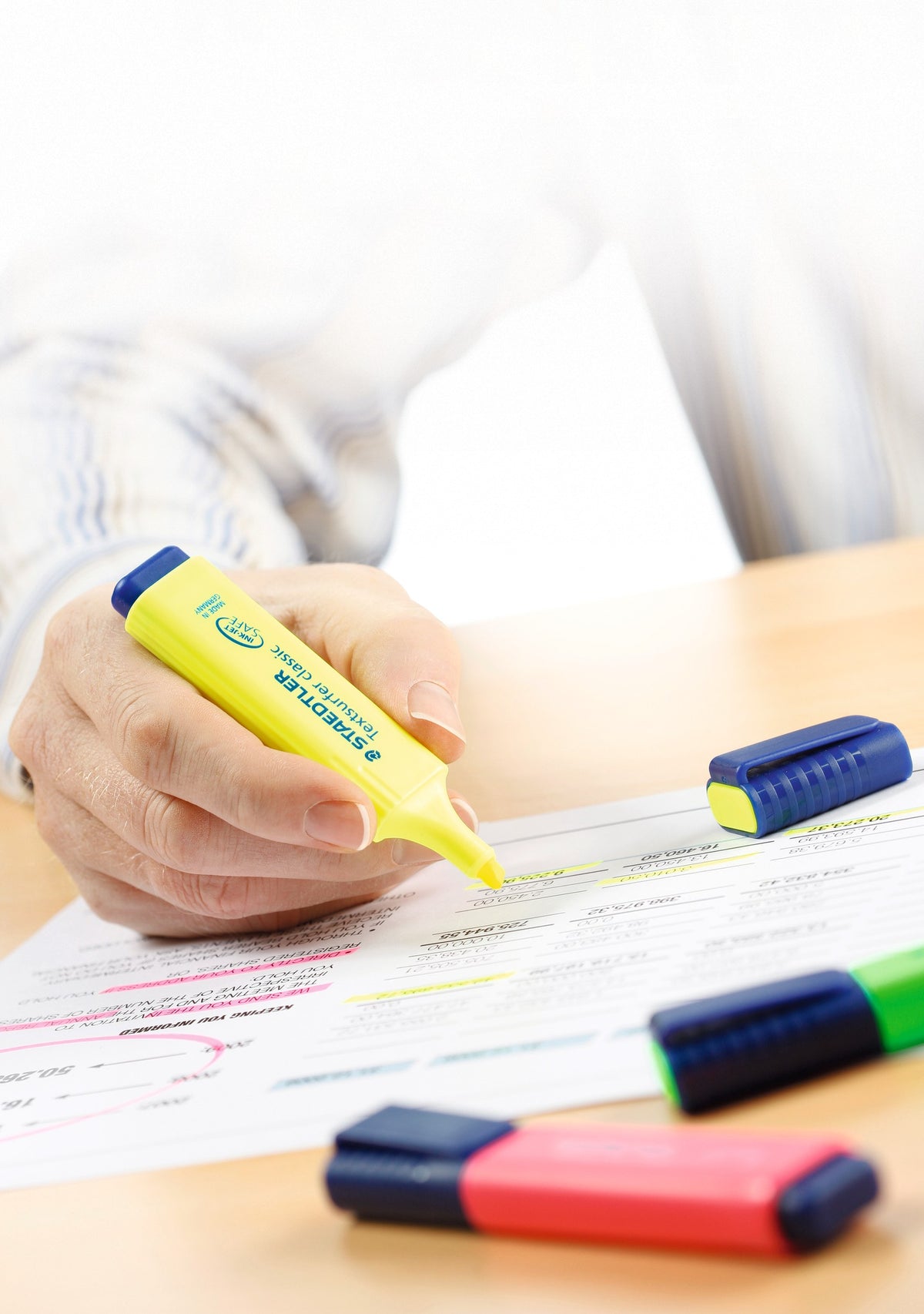 Desk scene of a hand highlighting paperwork with a yellow Textsurfer classic, capped pens lying nearby