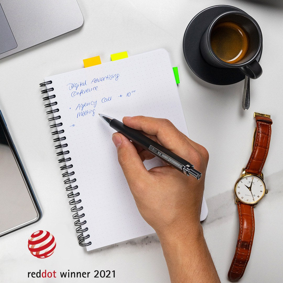 Hand writing in a spiral notebook with the dark grey triplus ballpoint beside a coffee cup, laptop and watch, reddot winner 2021 logo