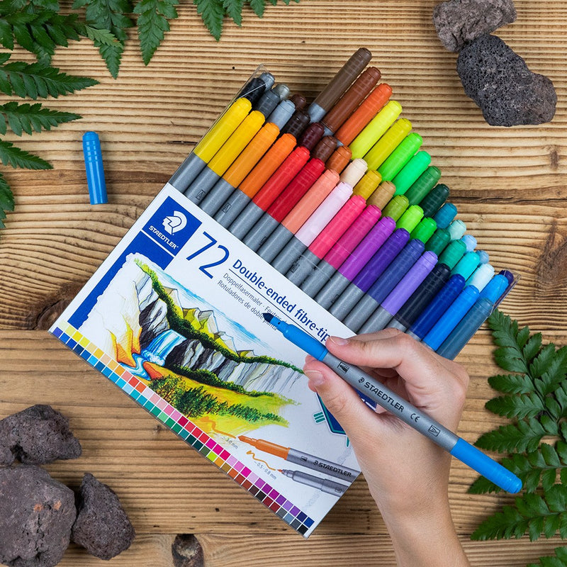 Staedtler photo of a hand drawing with a 3200 double-ended pen beside the open wallet of 72 on a timber table with ferns and stones