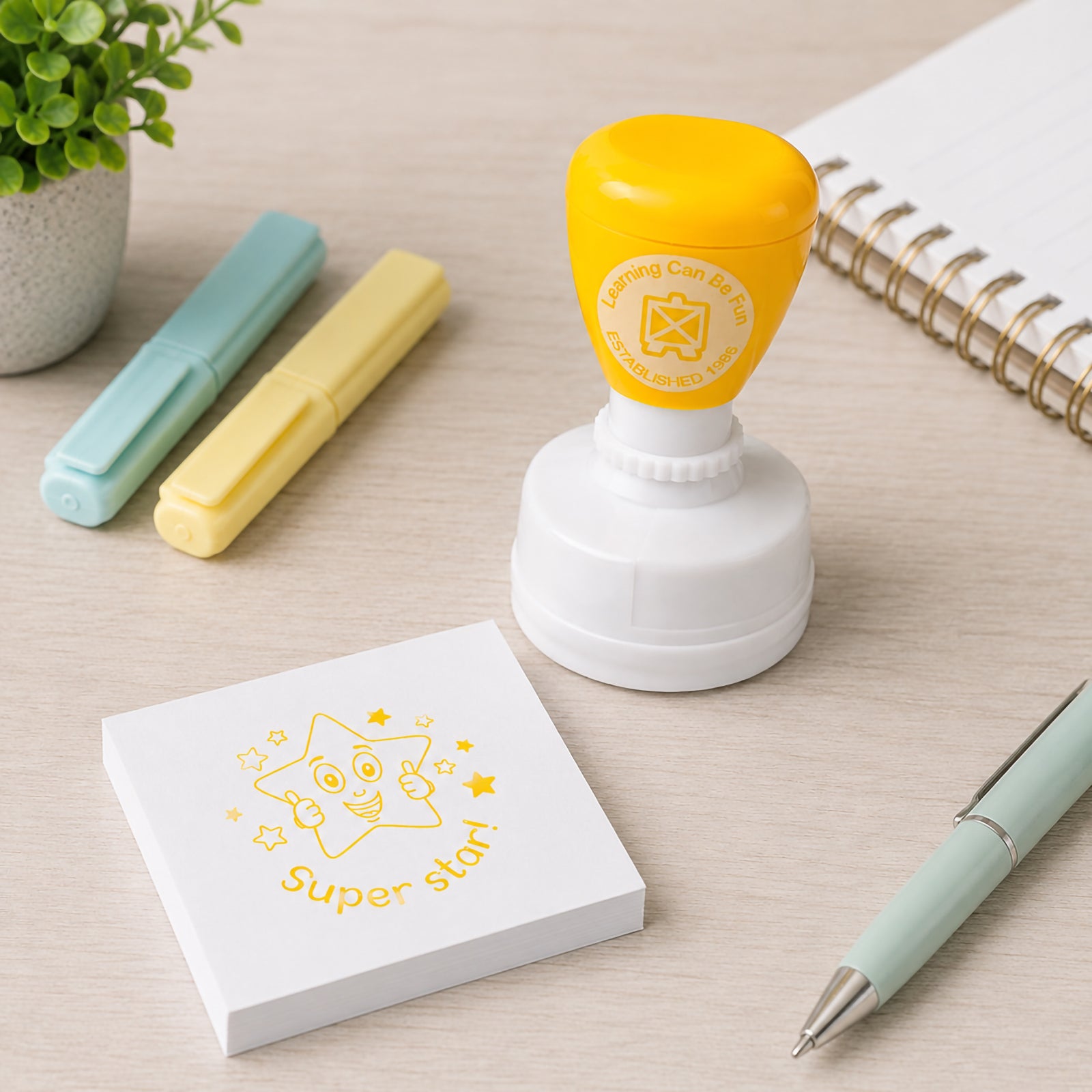 Super Star Merit Stamp displayed next to a memo pad, pastel markers and a pen on a light desk. The yellow stamp impression features a happy star with thumbs up, ideal for positive classroom feedback and daily learning rewards.