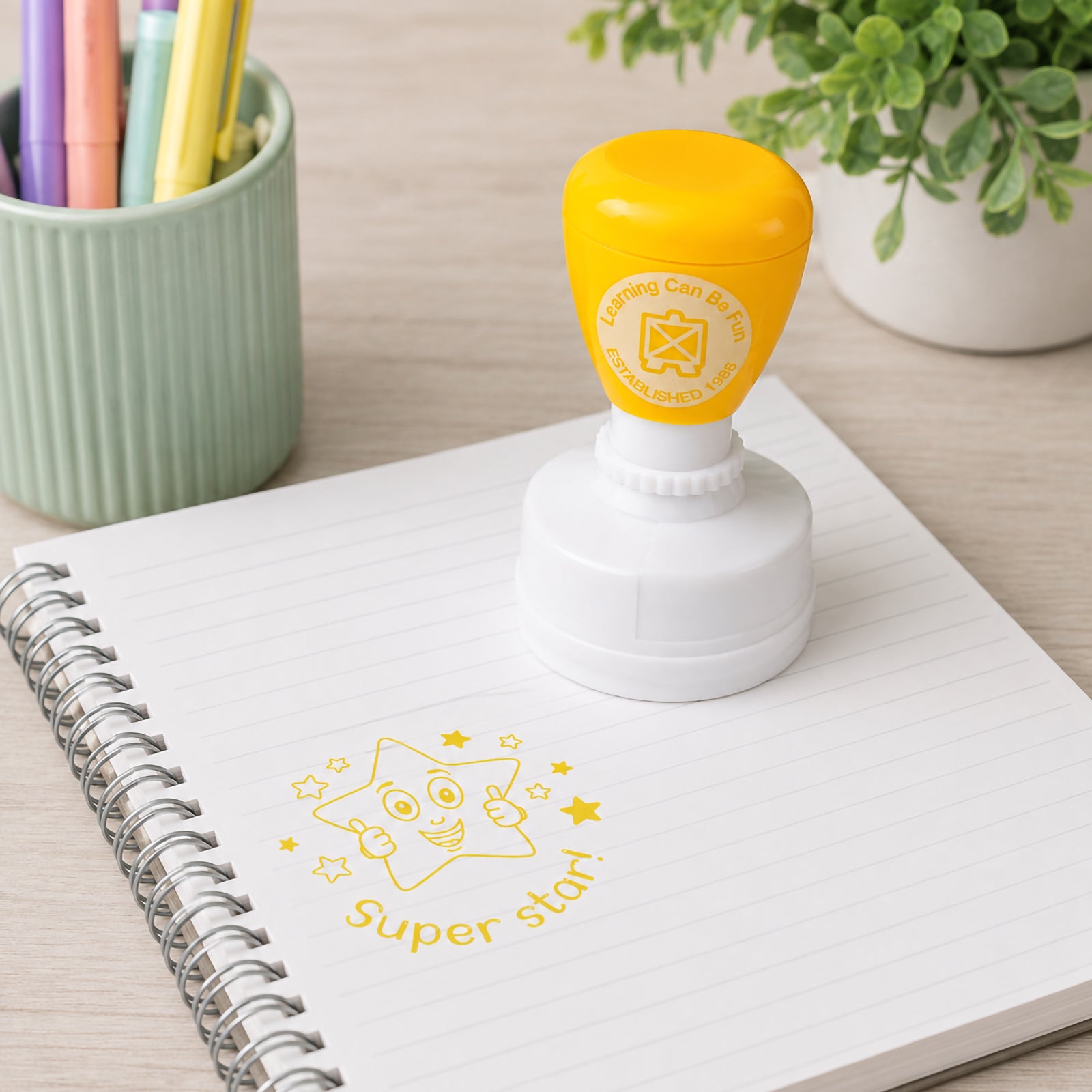 Learning Can Be Fun Super Star Merit Stamp shown on a lined notebook with a bright yellow smiling star impression. The self inking teacher stamp sits beside classroom stationery and plants, presenting a cheerful reward tool for recognising effort and student achievement.
