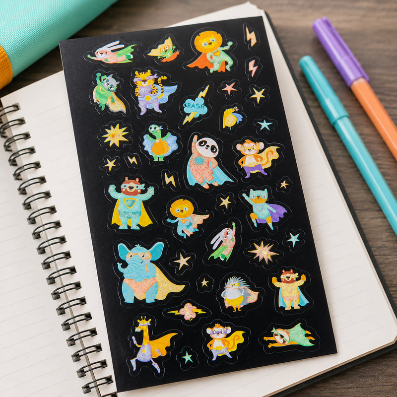 Superheroes glow sticker sheet displayed on an open notebook with colourful pens nearby. The collection showcases 37 vibrant animal hero characters, stars and lightning bolts designed for creative decorating and craft activities.