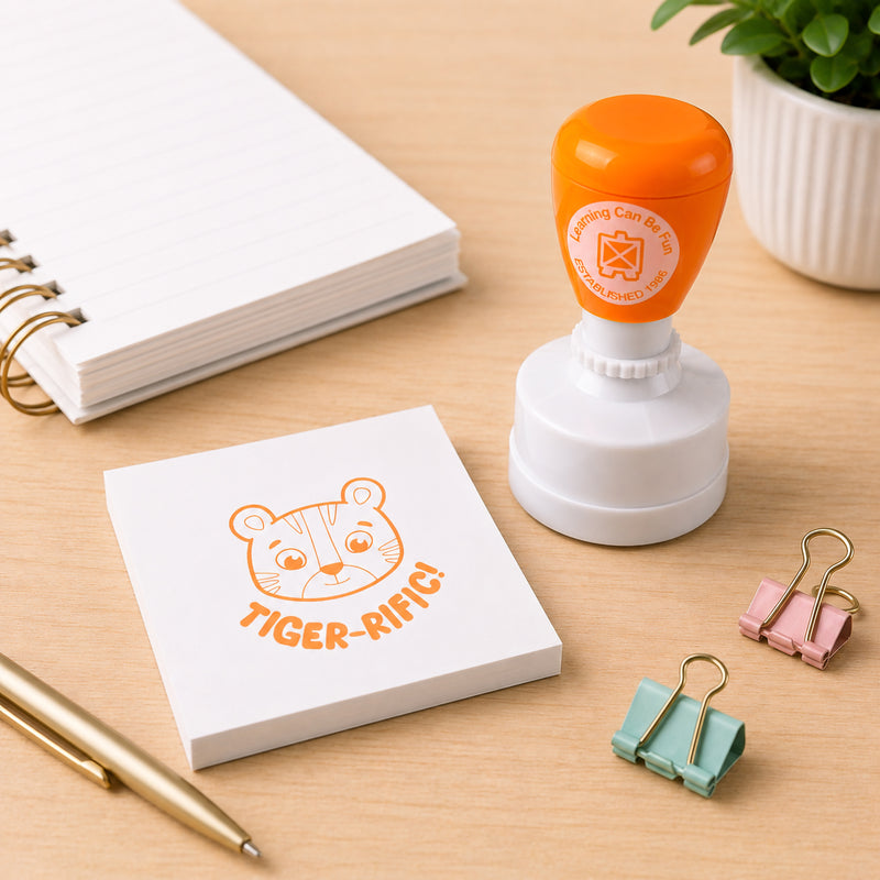 Learning Can Be Fun Tiger-rific Merit Stamp displayed on a classroom desk beside a memo pad featuring a bright tiger themed stamp impression. The orange self inking stamp is surrounded by stationery accessories, showcasing its use as a fun reward tool for recognising student achievement.