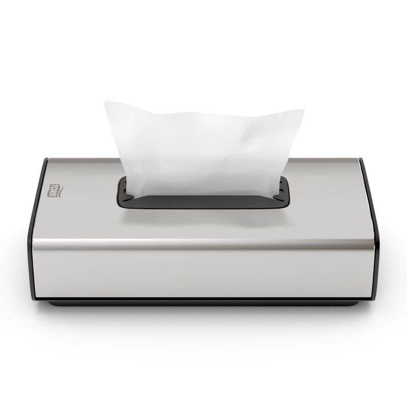 Brushed stainless steel Tork facial tissue dispenser in use, with a white tissue drawn up through the slot in the top face