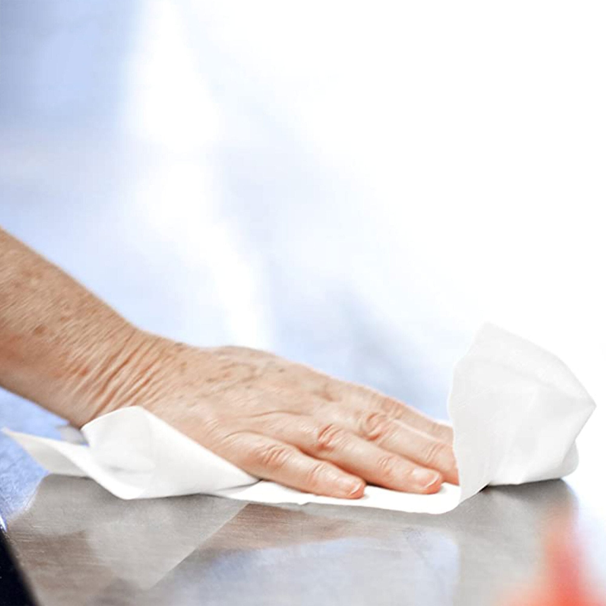 A hand pressing a crumpled sheet of white Tork Basic centrefeed wiping paper flat against a stainless steel bench top, wiping the surface clean, photographed against a soft pale blue background