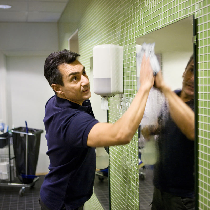 Cleaner in a navy polo shirt wiping a washroom mirror with a paper sheet, with a white Tork M1 mini centrefeed dispenser wall mounted on the green tiled wall beside him