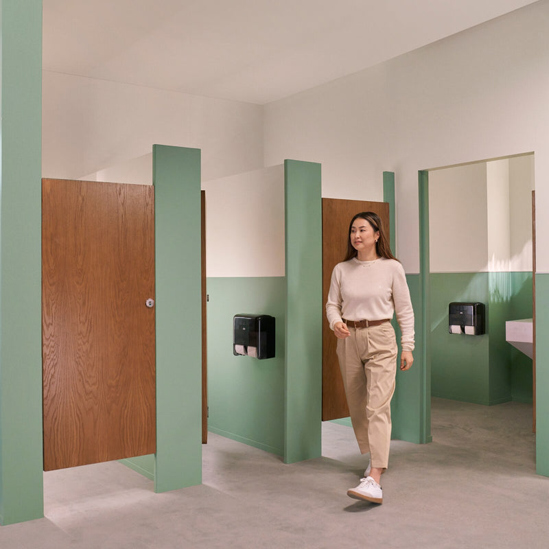 Tork T7 coreless toilet roll dispenser mounted on a tiled washroom wall inside a cubicle with a hand drawing a sheet from the dispensing slot.