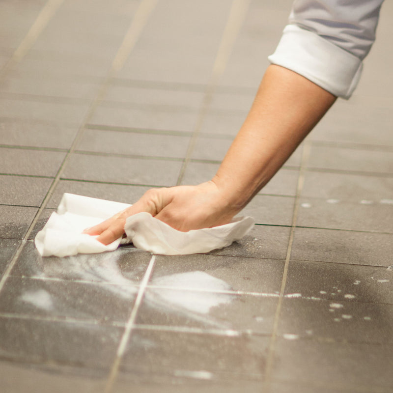 A person with a rolled up grey sleeve wipes spilled white liquid from grey tiled flooring using a crumpled sheet of white Tork centrefeed wiping paper, cleaned tiles visible behind the hand.