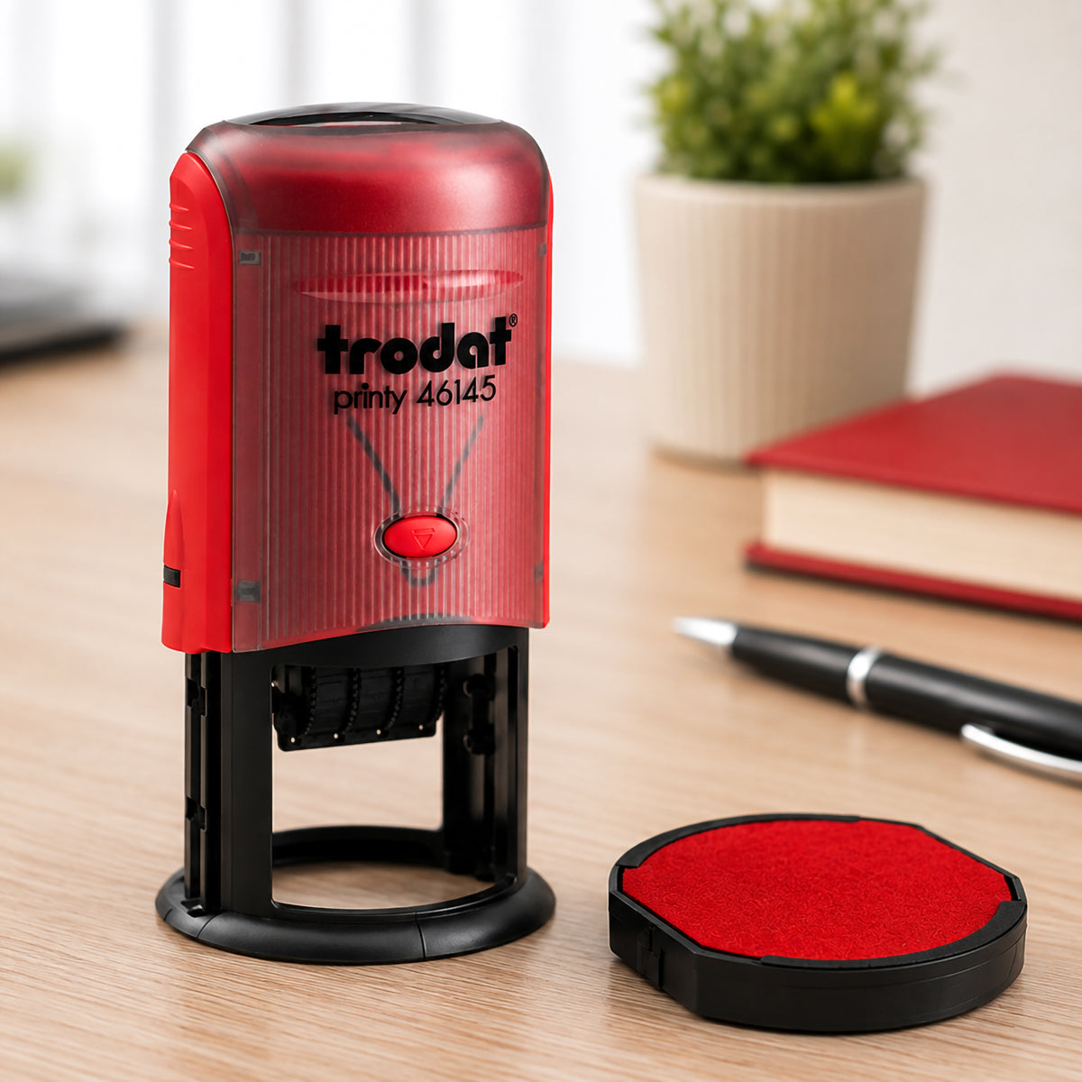 Close up of the Trodat Printy 46145 self inking stamp displayed alongside the genuine Trodat 6/46045 red replacement ink pad. The clean office workspace highlights the replacement pad used to maintain sharp, consistent stamp impressions.