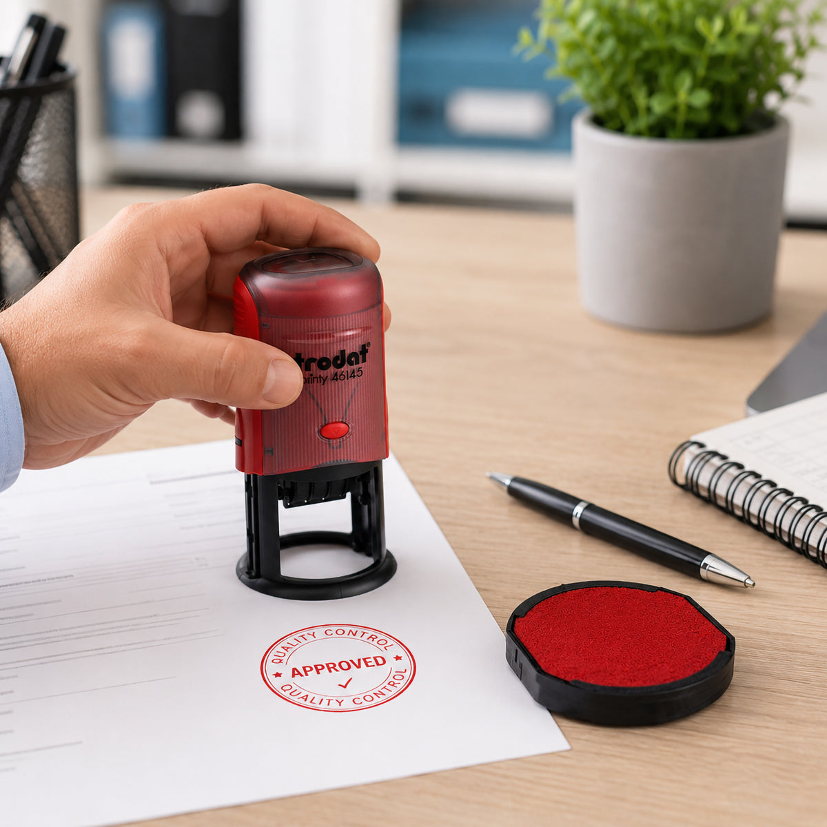 A user presses a Trodat Printy 46145 self inking stamp onto a business document, creating a crisp red Approved impression. The matching replacement red ink pad sits beside the stamp on a tidy office desk, demonstrating everyday professional document processing.