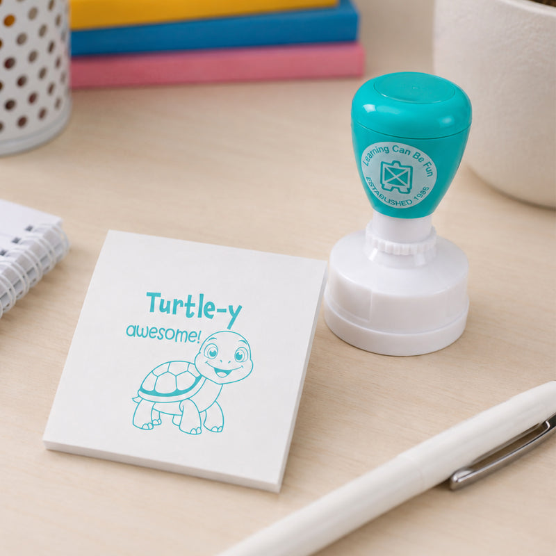 Self inking Turtle-y Awesome Merit Stamp positioned beside a memo pad with a clear turtle impression. The image showcases the compact design and suitability for rewarding classroom participation and learning progress.