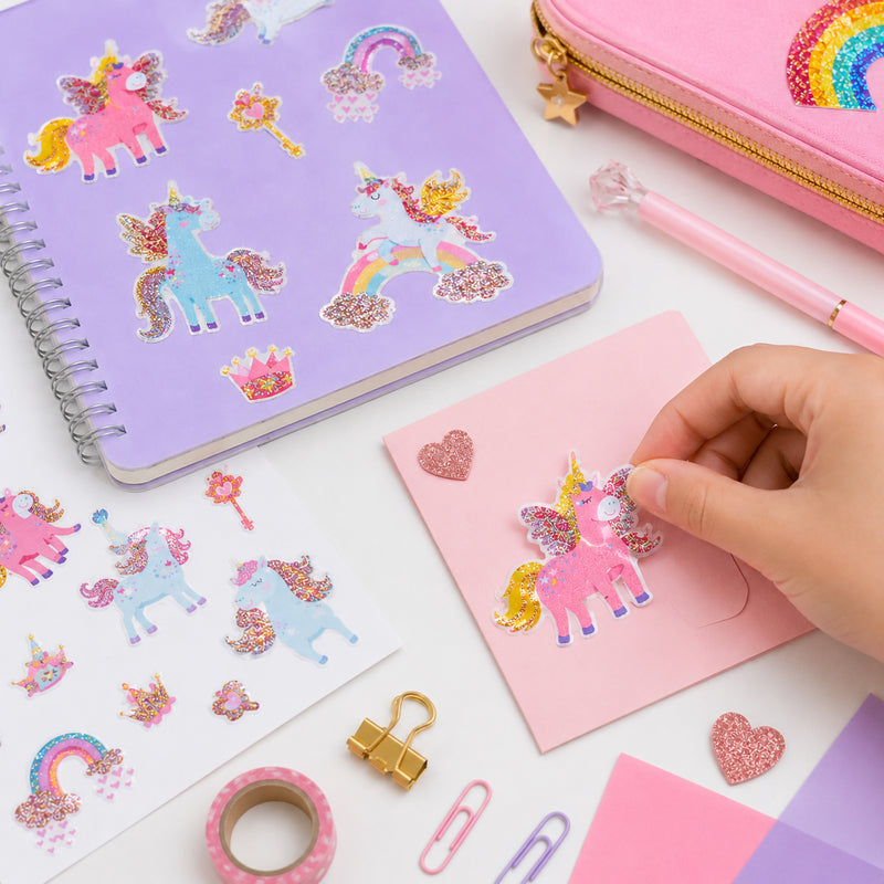 Child placing a glitter unicorn sticker onto a pink card surrounded by colourful stationery and craft accessories. The sparkling unicorn designs feature pastel pink, blue and gold colours with magical fantasy themed details.
