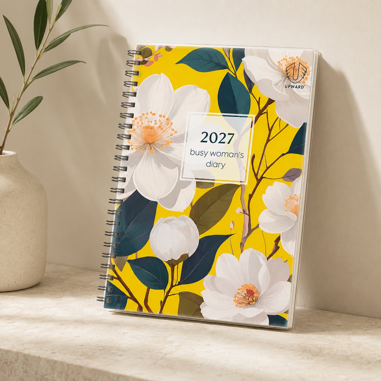 The Upward Busy Women's 2027 A5 Week To View Diary stands upright on a stone shelf with decorative greenery, showcasing the bright Bird Of Paradise inspired floral artwork, sturdy spiral binding and protective cover designed for everyday planning.