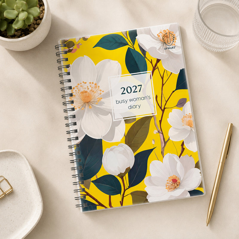 Top view of the Upward Busy Women's 2027 A5 Week To View Diary displayed beside a gold pen, glass of water and decorative plant on a light desktop, highlighting the colourful botanical cover and compact A5 weekly planning format.