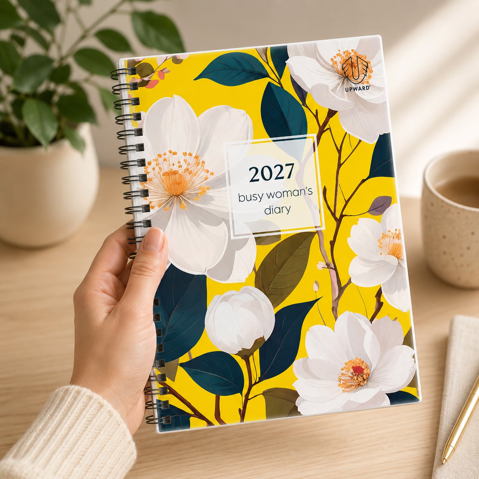 A person holds the Upward Busy Women's 2027 A5 Week To View Diary featuring a vibrant Bird Of Paradise inspired floral cover with bright yellow background, elegant white flowers, durable Wire O binding and protective clear cover, creating a stylish planner for everyday organisation.