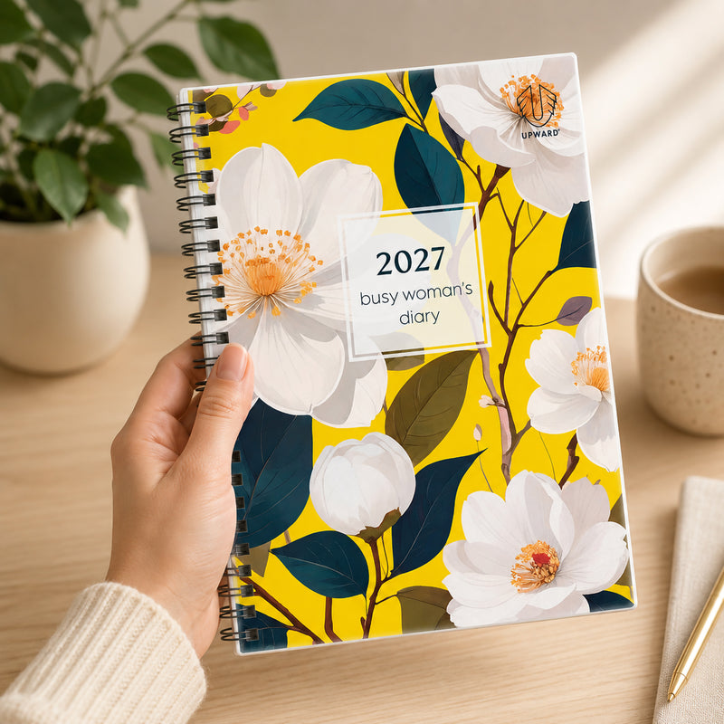 A person holds the Upward Busy Women's 2027 A5 Week To View Diary featuring a vibrant Bird Of Paradise inspired floral cover with bright yellow background, elegant white flowers, durable Wire O binding and protective clear cover, creating a stylish planner for everyday organisation.