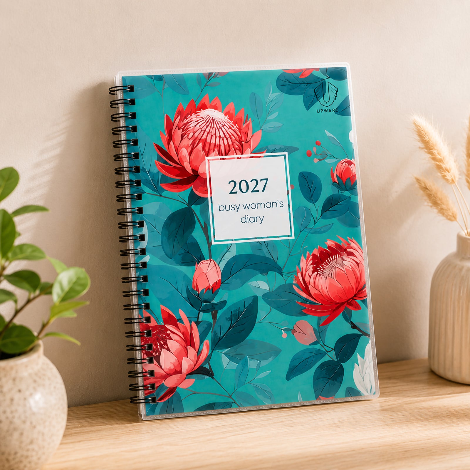 The Upward Busy Women's 2027 A5 Week To View Diary stands upright on a desk surrounded by decorative plants and home office accessories, highlighting the vivid floral artwork, sturdy spiral binding and protective clear cover designed for everyday planning.