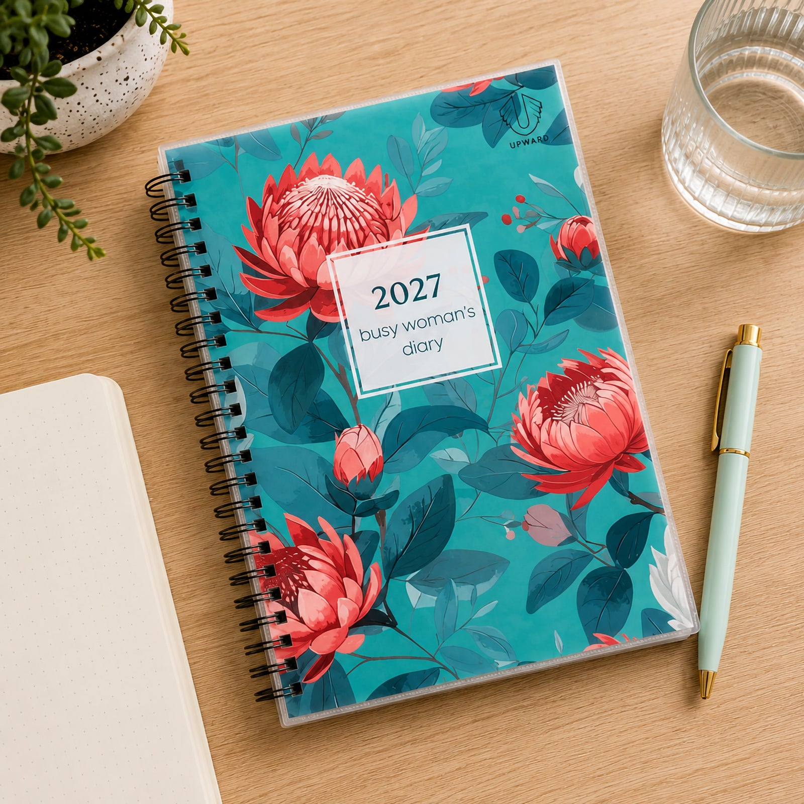Top view of the Upward Busy Women's 2027 A5 Week To View Diary displayed beside a notebook, pen, glass of water and indoor plant on a timber desk. The colourful botanical cover and compact A5 format create an attractive and practical weekly planning solution.