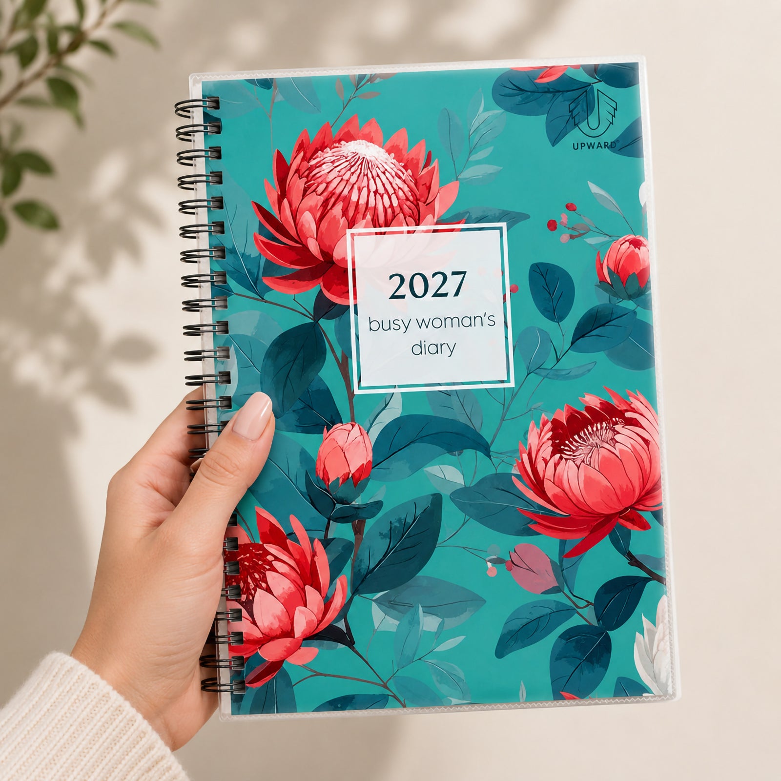 A hand holds the Upward Busy Women's 2027 A5 Week To View Diary featuring a vibrant Golden Wattle inspired botanical cover with colourful floral artwork, durable Wire O binding and protective clear cover, presenting a stylish planner suitable for everyday organisation.
