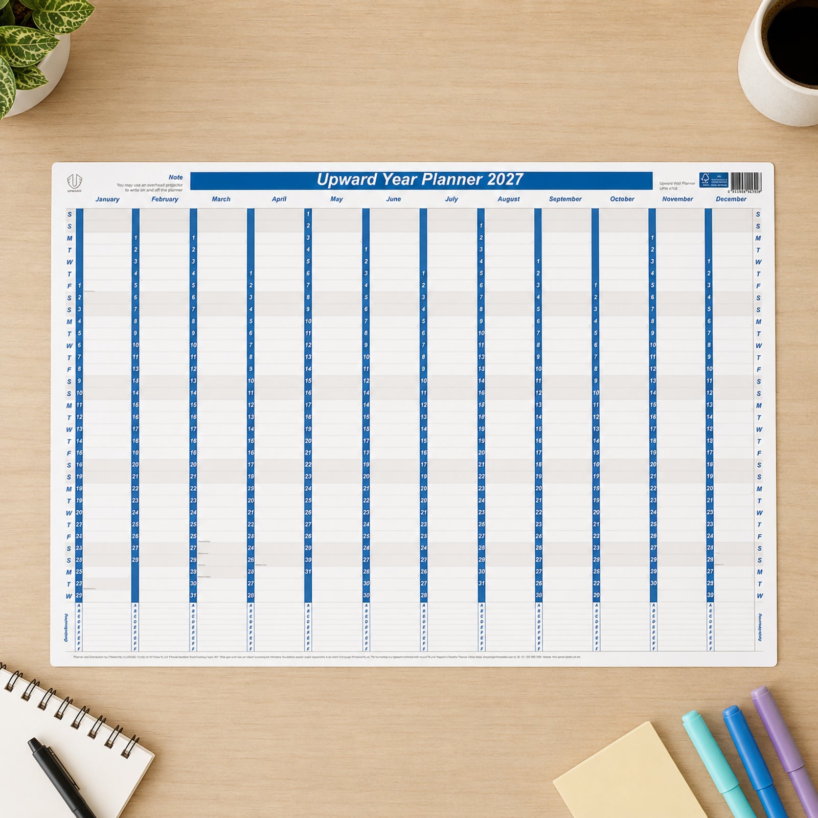 Top view of the Upward 2027 Laminated Year Planner positioned on a tidy workspace with notebooks, coloured markers, sticky notes and coffee. The reusable wall planner presents the full twelve month calendar in a compact layout suitable for office, study or home planning.