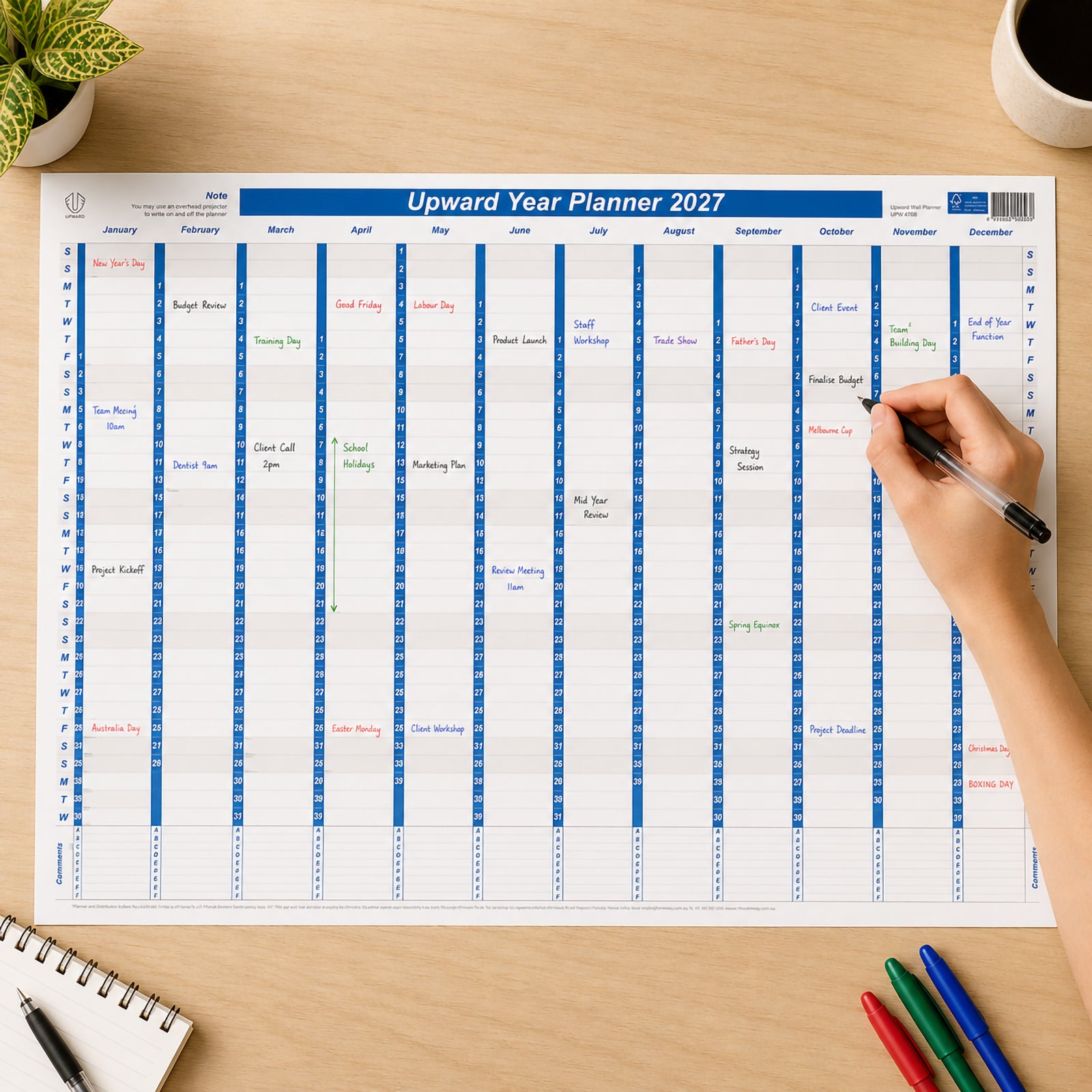 The Upward 2027 Laminated Year Planner is displayed on a desk while a hand writes upcoming meetings and deadlines onto the reusable write on wipe off surface. The planner shows the complete year at a glance with Australian public holidays, demonstrating how easily schedules can be updated throughout the year.