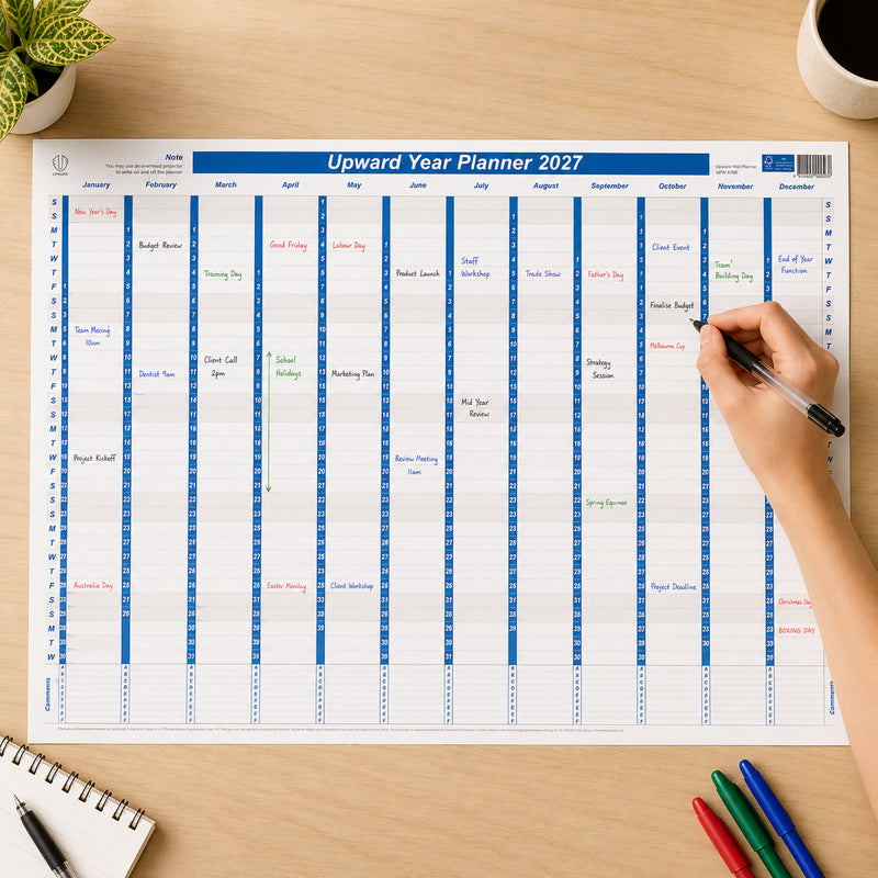 The Upward 2027 Laminated Year Planner is displayed on a desk while a hand writes upcoming meetings and deadlines onto the reusable write on wipe off surface. The planner shows the complete year at a glance with Australian public holidays, demonstrating how easily schedules can be updated throughout the year.