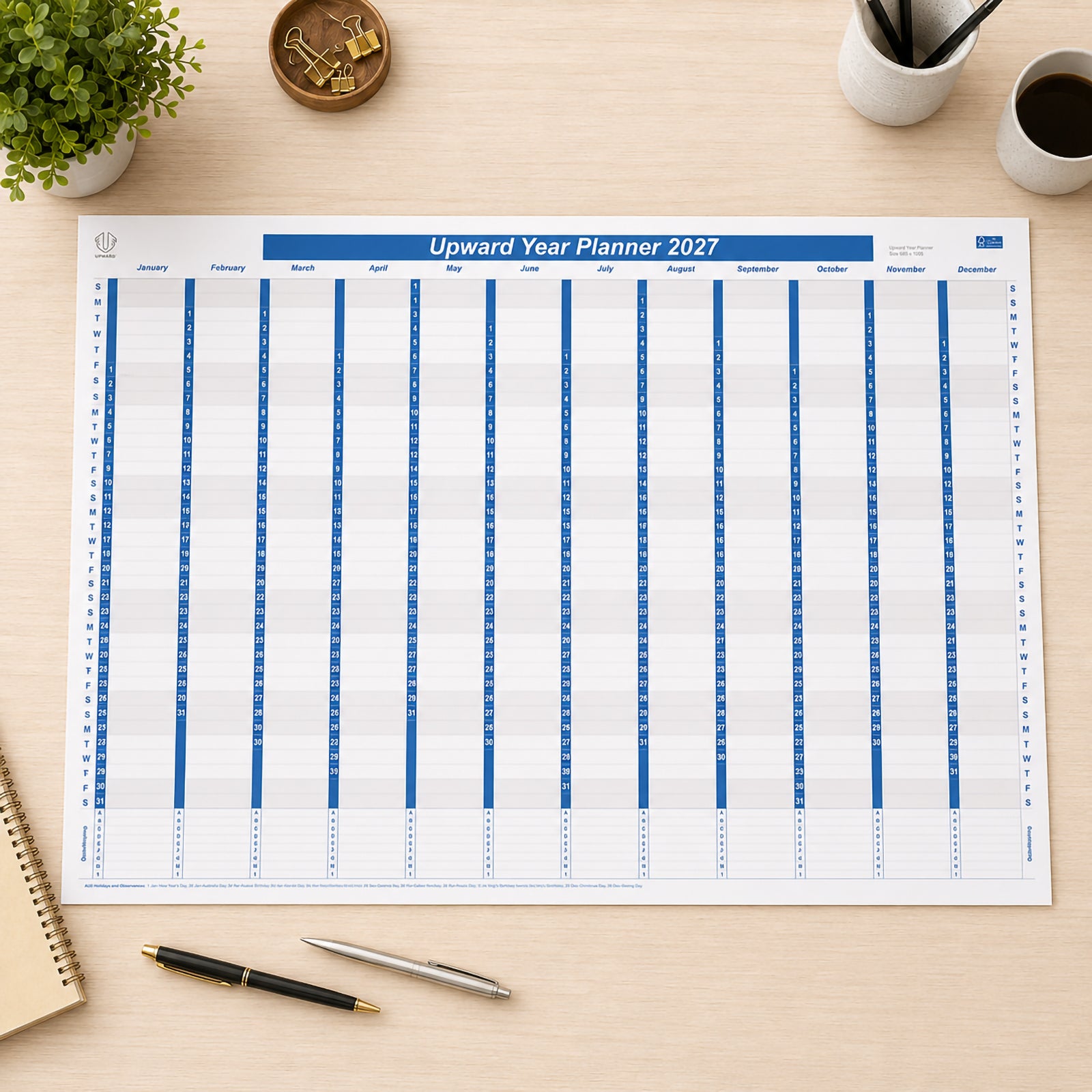 Top view of the Upward 2027 Year Planner laid flat on a clean desk surrounded by office stationery, notebooks and coffee. The large 1005 x 685mm paper planner provides a complete year at a glance, making it ideal for organising business projects, family events and long term schedules.