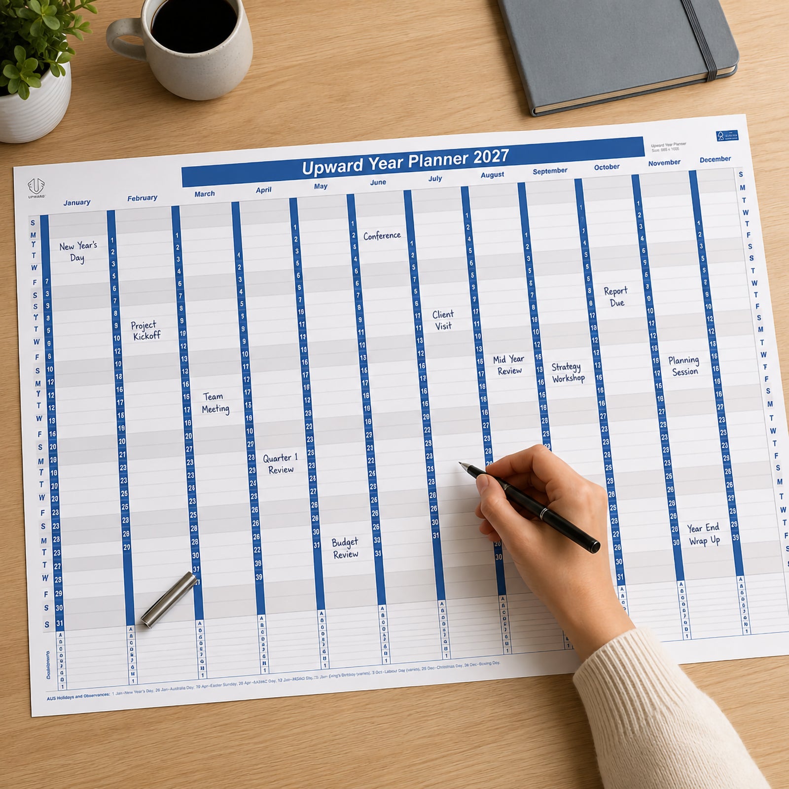 The Upward 2027 Year Planner is displayed on a wooden desk while a hand writes upcoming appointments onto the large year to view calendar. The planner clearly displays all twelve months with Australian public holidays, demonstrating its practical use for scheduling meetings, project deadlines and annual planning in a professional workspace.